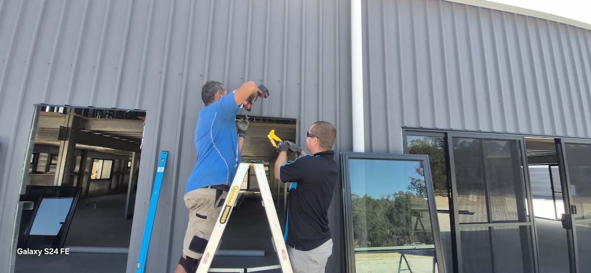 Two men work together outside a gray industrial building—one stands on a ladder using a drill while the other hands him a tool, showcasing Construction and Maintenance Solutions. A large window reflects nearby trees.