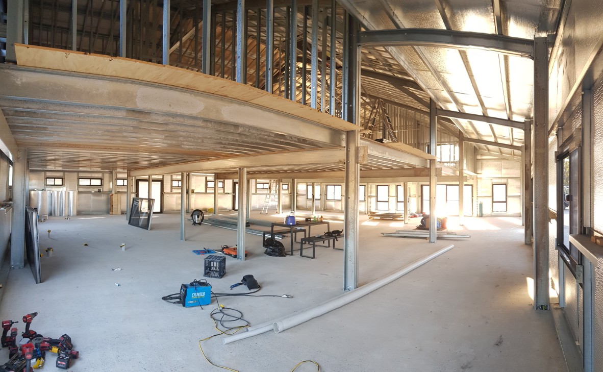 Spacious interior of a building under construction, with exposed metal framing, tools, and pipes scattered on the concrete floor—showcasing Construction and Maintenance Solutions in action across unfinished upper and lower levels.