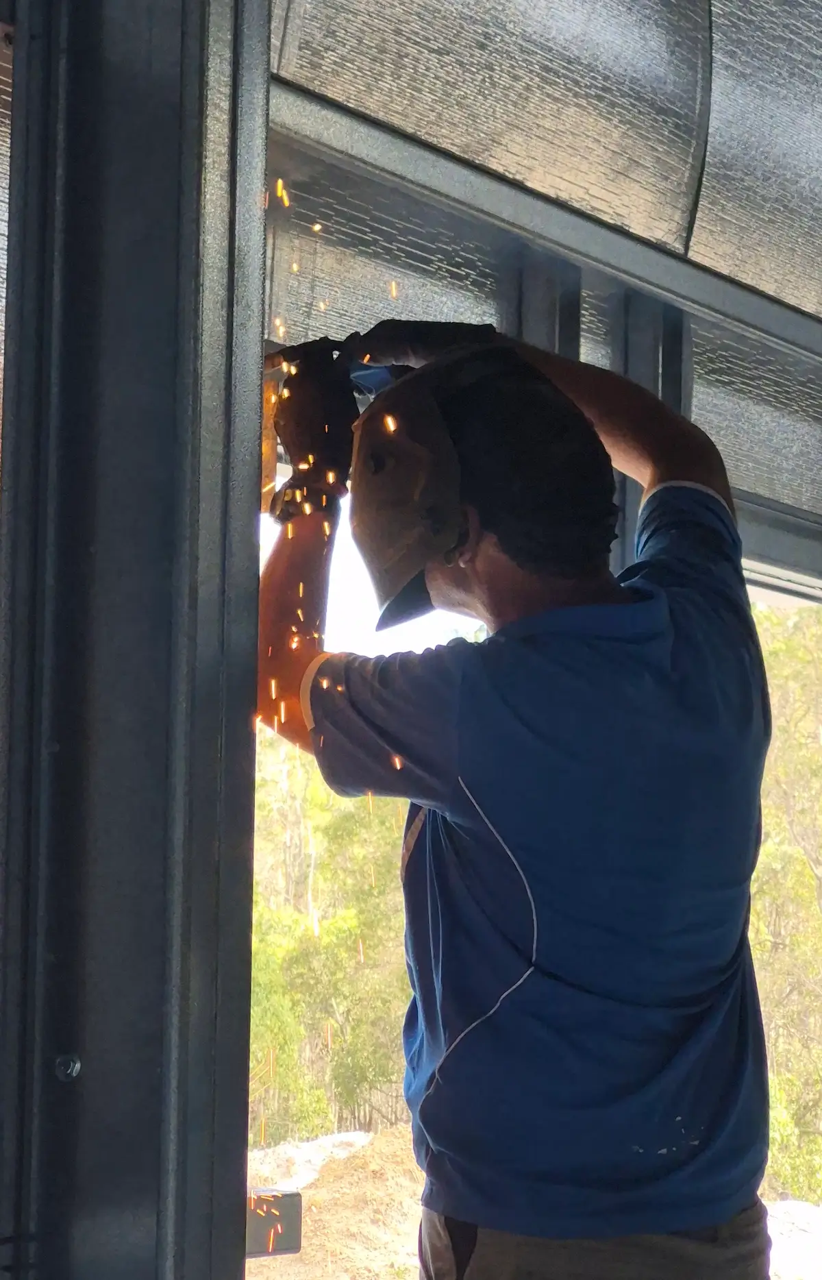 A person wearing a cap and gloves welds a metal frame indoors, sparks flying as sunlight streams through windows. Construction and Maintenance Solutions shine through, with trees illuminated outside in the background.
