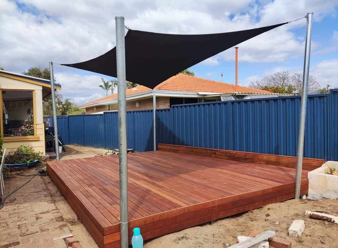 A backyard features a raised wooden deck with metal posts, expertly crafted by Construction and Maintenance Solutions, supporting a black shade sail overhead. The area is surrounded by a blue steel fence, with neighboring houses and a partly cloudy sky in the background.