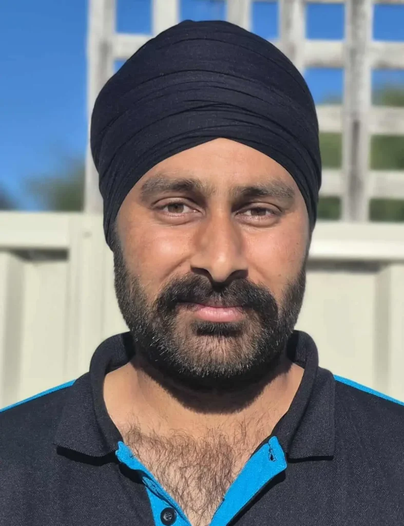 A man with a black turban and beard stands outdoors in sunlight, wearing a black collared shirt with blue trim, in front of a white fence and lattice.