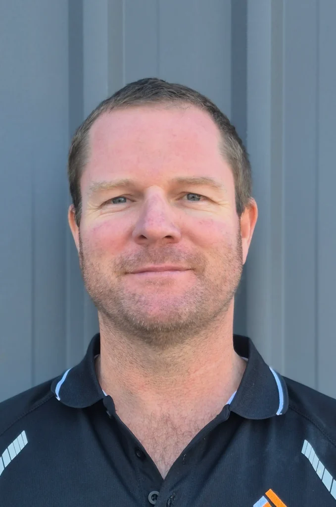 A man with short brown hair and light facial hair is wearing a black collared shirt with white trim, standing in front of a gray metal background, and smiling slightly at the camera.