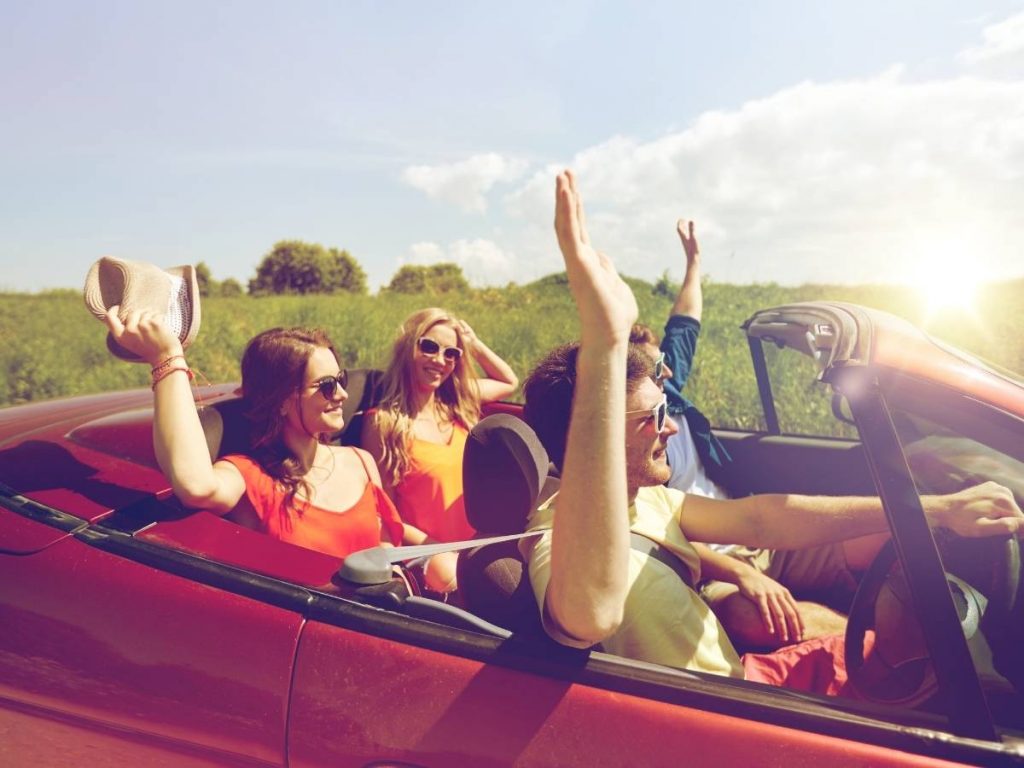 Four friends ride in a red convertible with the top down on a sunny day, smiling and wearing sunglasses as they drive through green countryside—showing that fun adventures and reliable Construction and Maintenance Solutions both create smooth journeys.