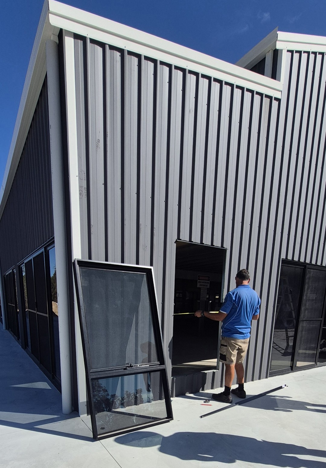 A person in a blue shirt and shorts installs or repairs a window on the side of a modern gray metal building, showcasing Construction and Maintenance Solutions, with a window frame resting nearby on the concrete surface.