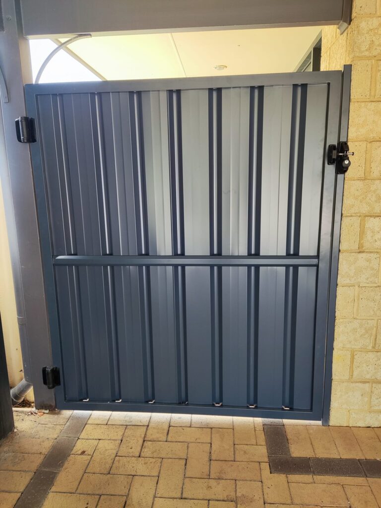 A blue metal gate with vertical grooves and a lock on the right side, set between a beige brick wall and a metal post, showcases quality Construction and Maintenance Solutions, complemented by a tiled floor beneath.