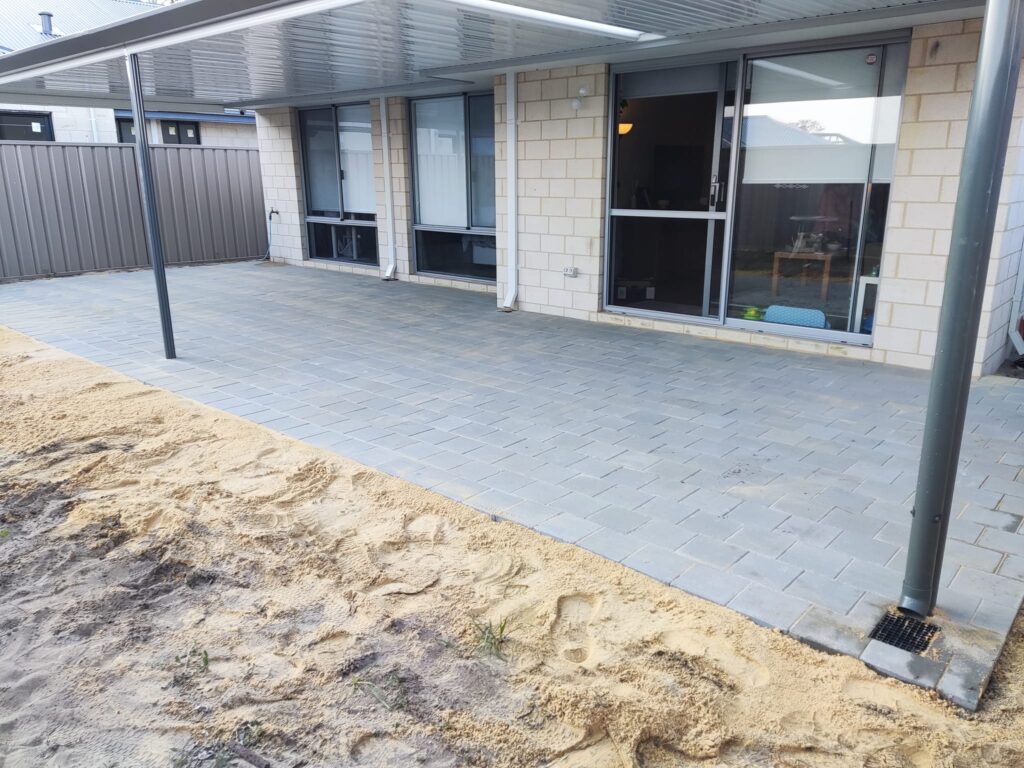 A newly paved patio area with light gray bricks next to a house, featuring large windows, sliding doors, and a covered roof with metal poles—showcasing quality Construction and Maintenance Solutions against the backdrop of bare sandy ground.