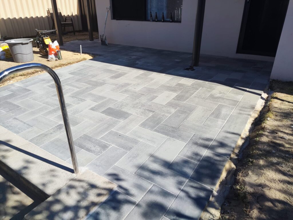 A newly paved patio with grey rectangular tiles in a herringbone pattern showcases expert Construction and Maintenance Solutions, set beside a building with tools, a bucket, and unfinished edges. A metal handrail and shadows appear in the foreground.