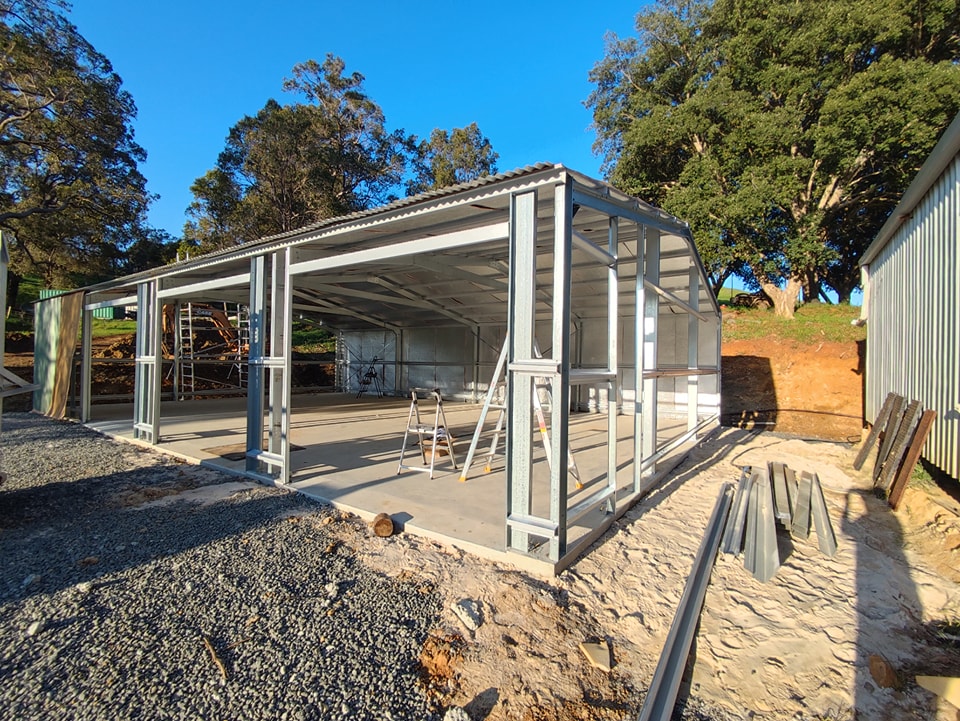 A partially constructed metal-framed building stands on a concrete foundation, surrounded by construction materials and trees under a bright blue sky, showcasing expert Construction and Maintenance Solutions. A ladder and scaffolding are visible inside and nearby.