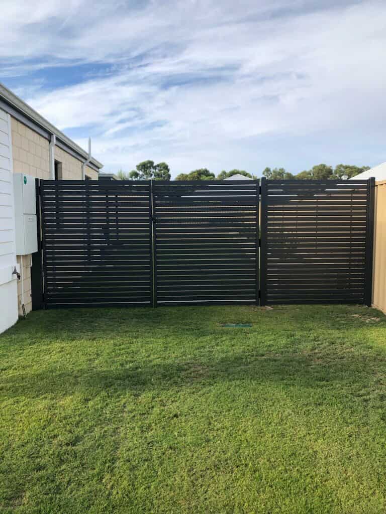 A modern, dark-colored slatted metal gate—designed with Construction and Maintenance Solutions in mind—stands between two buildings, enclosing a green lawn area under a partly cloudy sky.