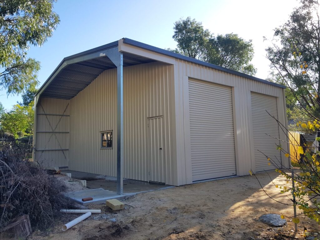 A beige metal shed with two large roller doors, a small window, and an open side section. Ideal for Construction and Maintenance Solutions, it sits on dirt ground among pipes, construction materials, and is surrounded by trees and bushes.