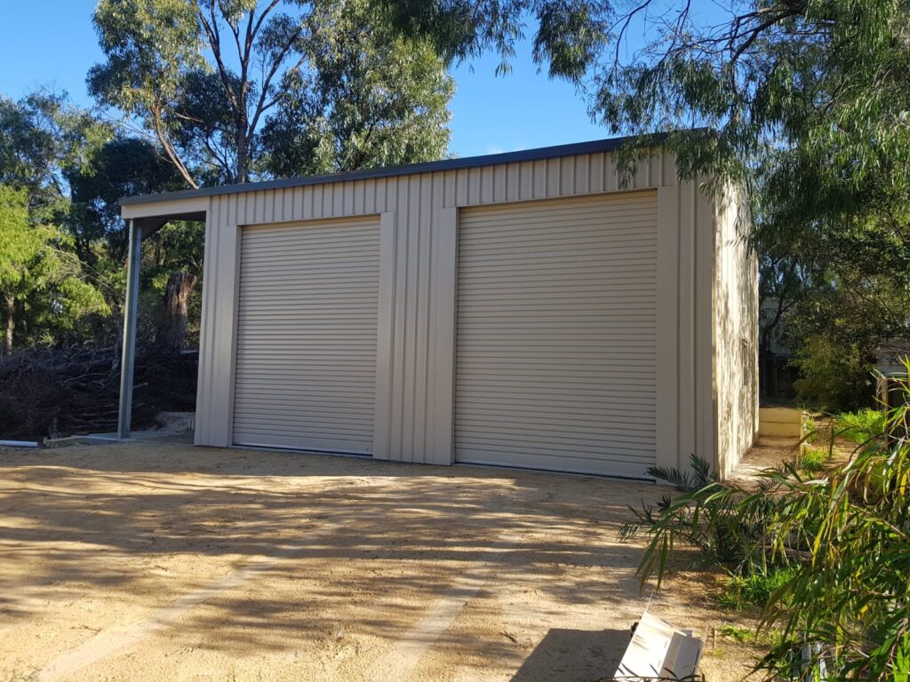 A light-colored metal shed with two large closed roller doors stands on a sandy patch, surrounded by trees and greenery—an ideal example of reliable construction and maintenance solutions in a rural outdoor setting.