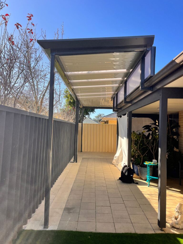 A modern outdoor patio with a transparent roof attached to a house, supported by dark metal posts. The area features expert Construction and Maintenance Solutions, with tiled paving, partial shade, stylish outdoor furniture, and nearby plants.