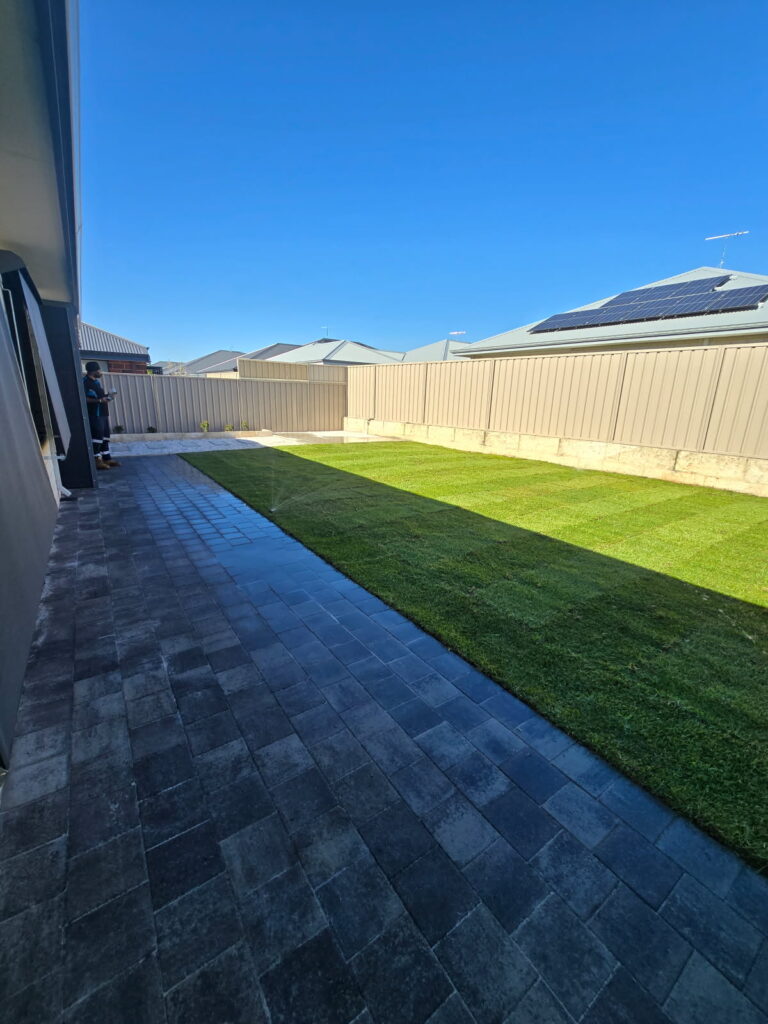A backyard with a neatly mowed rectangular lawn bordered by grey pavers and beige fencing, showcasing expert Construction and Maintenance Solutions. A person stands near the fence on the left. The sky is clear and blue.