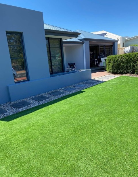 A modern house with blue-gray exterior walls, large windows, and a covered patio with chairs showcases expertly designed Construction and Maintenance Solutions. A green lawn and stone pathway bordered by white decorative rocks complete the sunny scene.