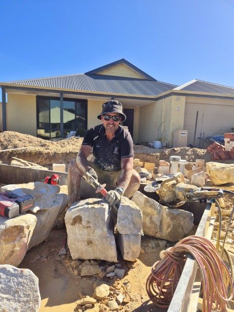 A man wearing sunglasses, a bucket hat, and gloves splits large rocks with a hammer outside a house under construction, showcasing hands-on Construction and Maintenance Solutions amid scattered materials and tools on a sunny day.