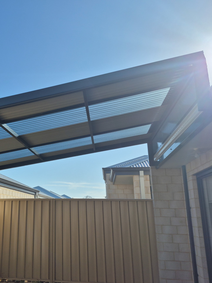 A clear corrugated patio roof of high-quality construction is attached to a beige brick house, with sunlight shining through. A beige metal fence and neighboring houses are seen in the background beneath a blue sky.