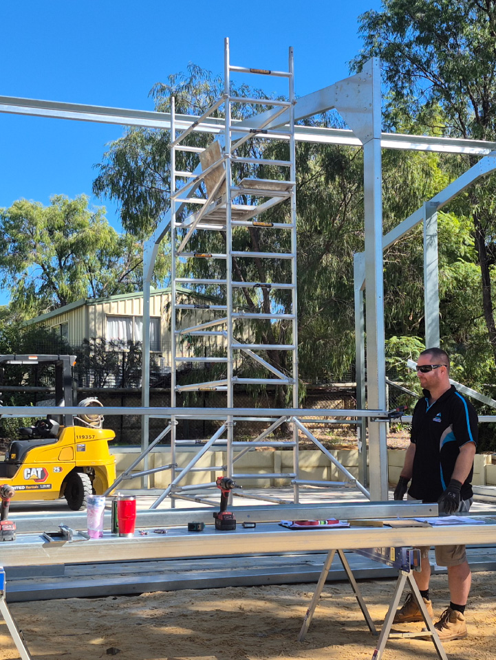 A man in sunglasses stands by a worktable with tools and drinks in front of a high-quality construction site featuring a partially built metal structure. A metal ladder, small yellow construction vehicle, and surrounding trees complete the scene.