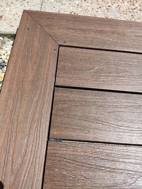 Close-up of a wooden or wood-look deck with diagonal edge trim and parallel planks, highlighting the wood grain texture. Showcasing quality Construction and Maintenance Solutions, with pebbles and concrete tiles visible in the background.