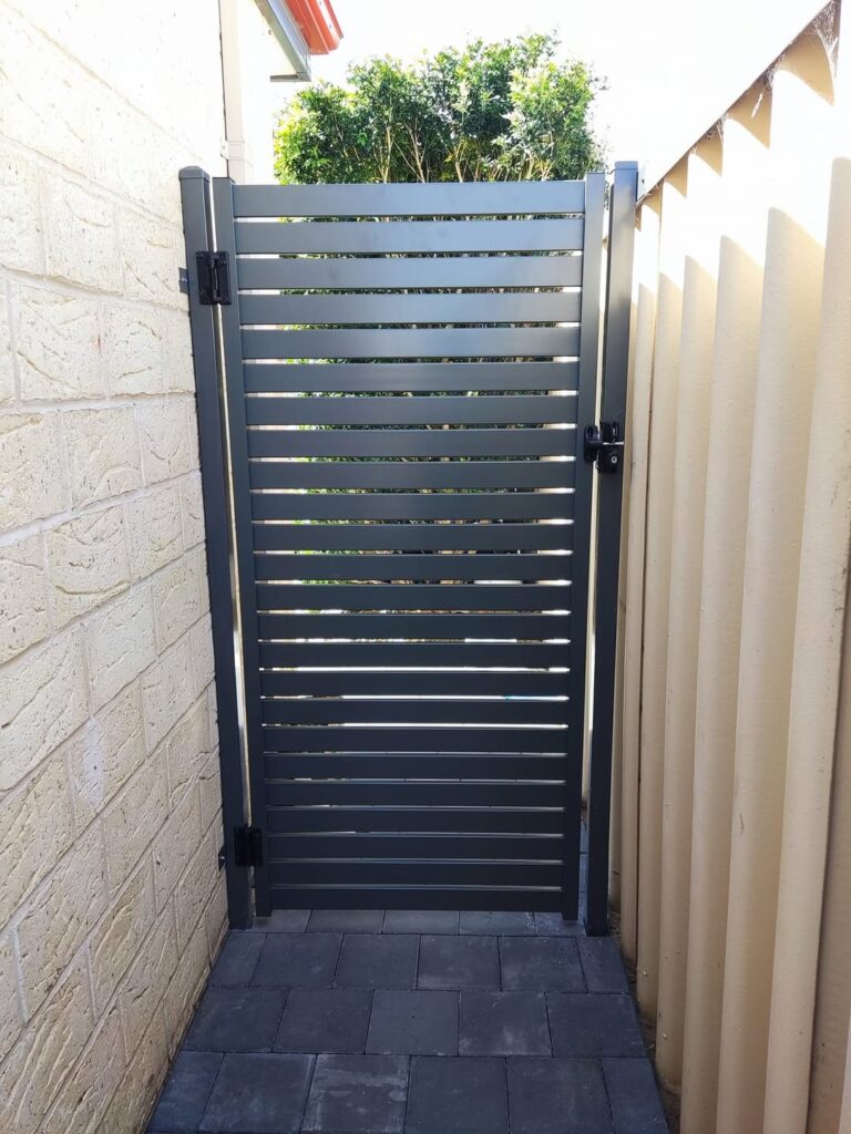 A modern grey metal slat gate by Construction and Maintenance Solutions is installed between a cream brick wall and a beige fence, leading to a paved pathway with greenery visible through the gate’s horizontal slats.